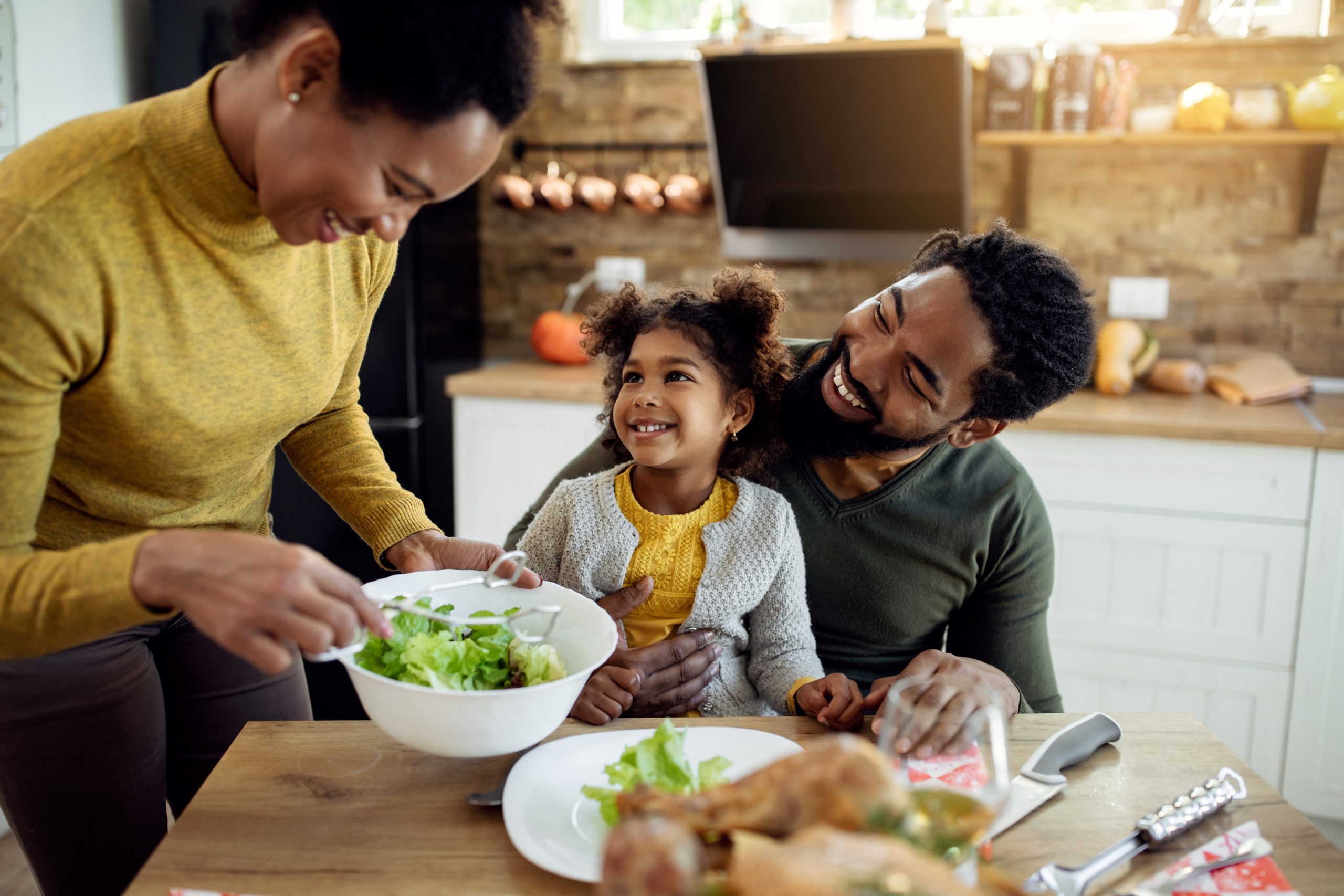 Family eating a meal together. Daughter sits on father's lap while mother serves salad.